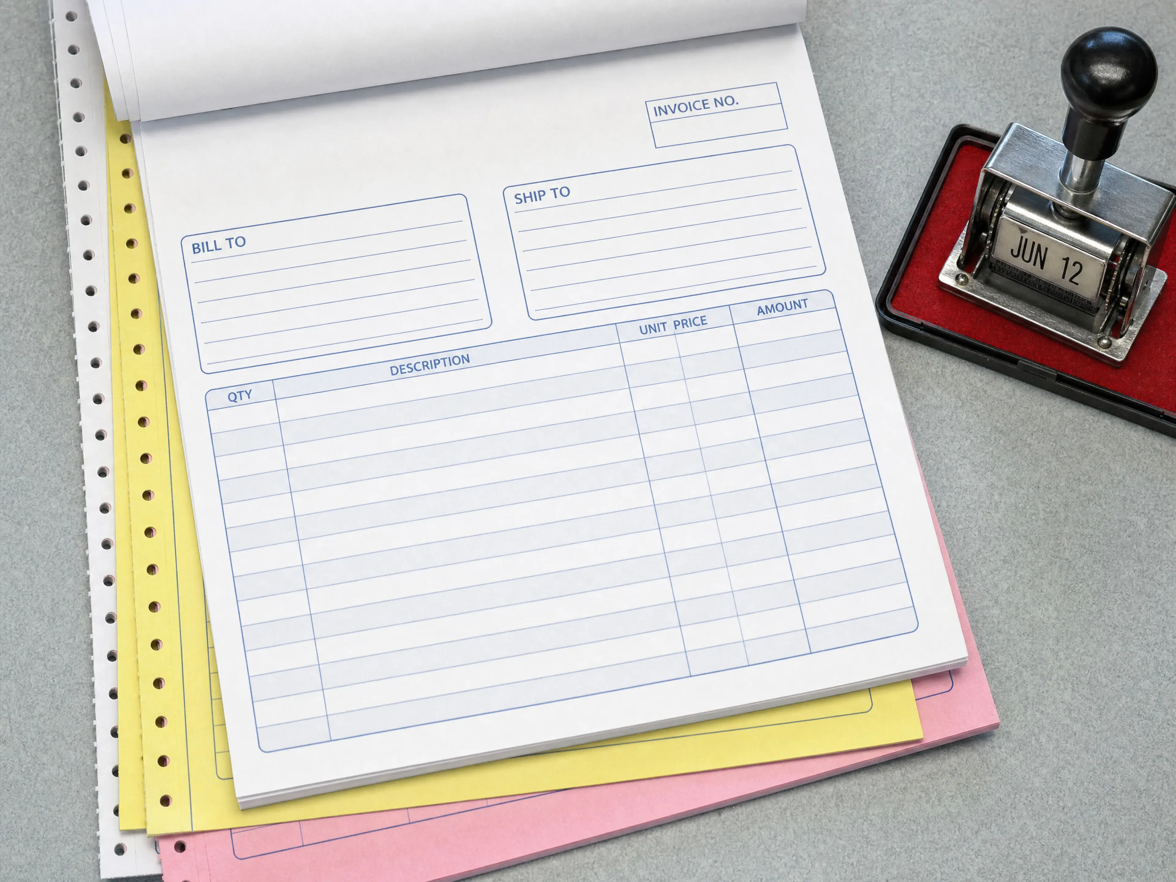 A fanned carbonless triplicate invoice pad on a fluorescent desk, white, canary, and pink sheets with tractor-feed holes, beside a steel date stamp on a red ink pad.
